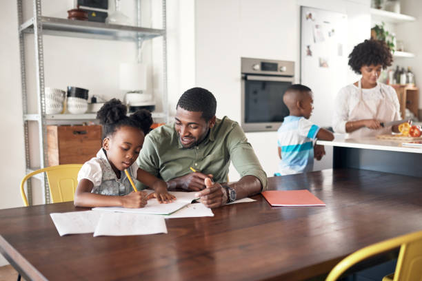 Shot of young man helping his daughter with her homework while her mother cooks in the background